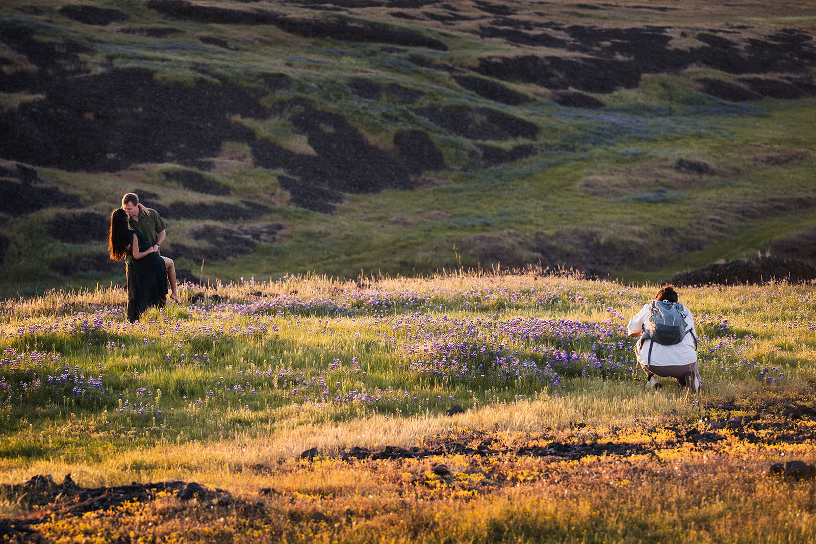 A photographer with a backpack crouches low in a meadow of golden grass and purple wildflowers at golden hour, camera raised toward a couple embracing nearby.