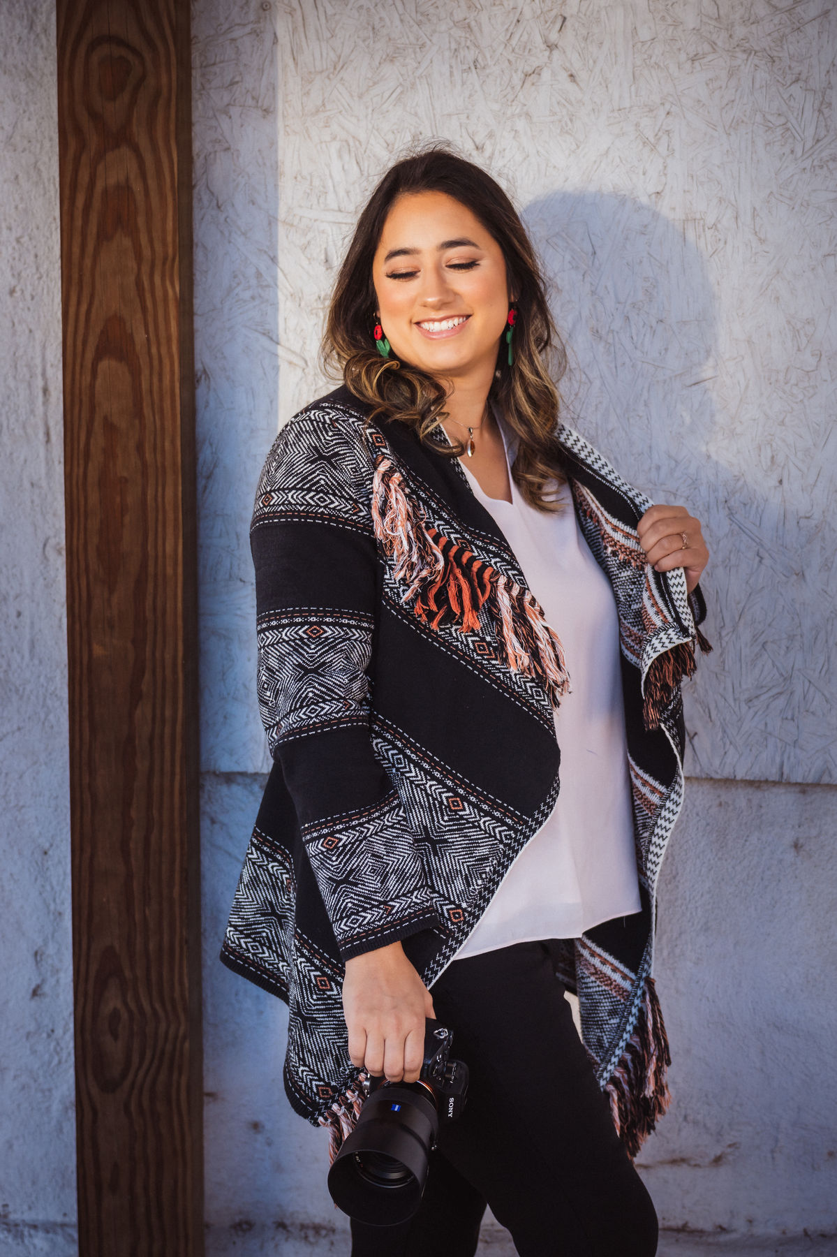 Maya Isabella smiling and looking down, wearing a black and white patterned fringed cardigan, holding a camera with a large lens at her side against a pale textured wall.